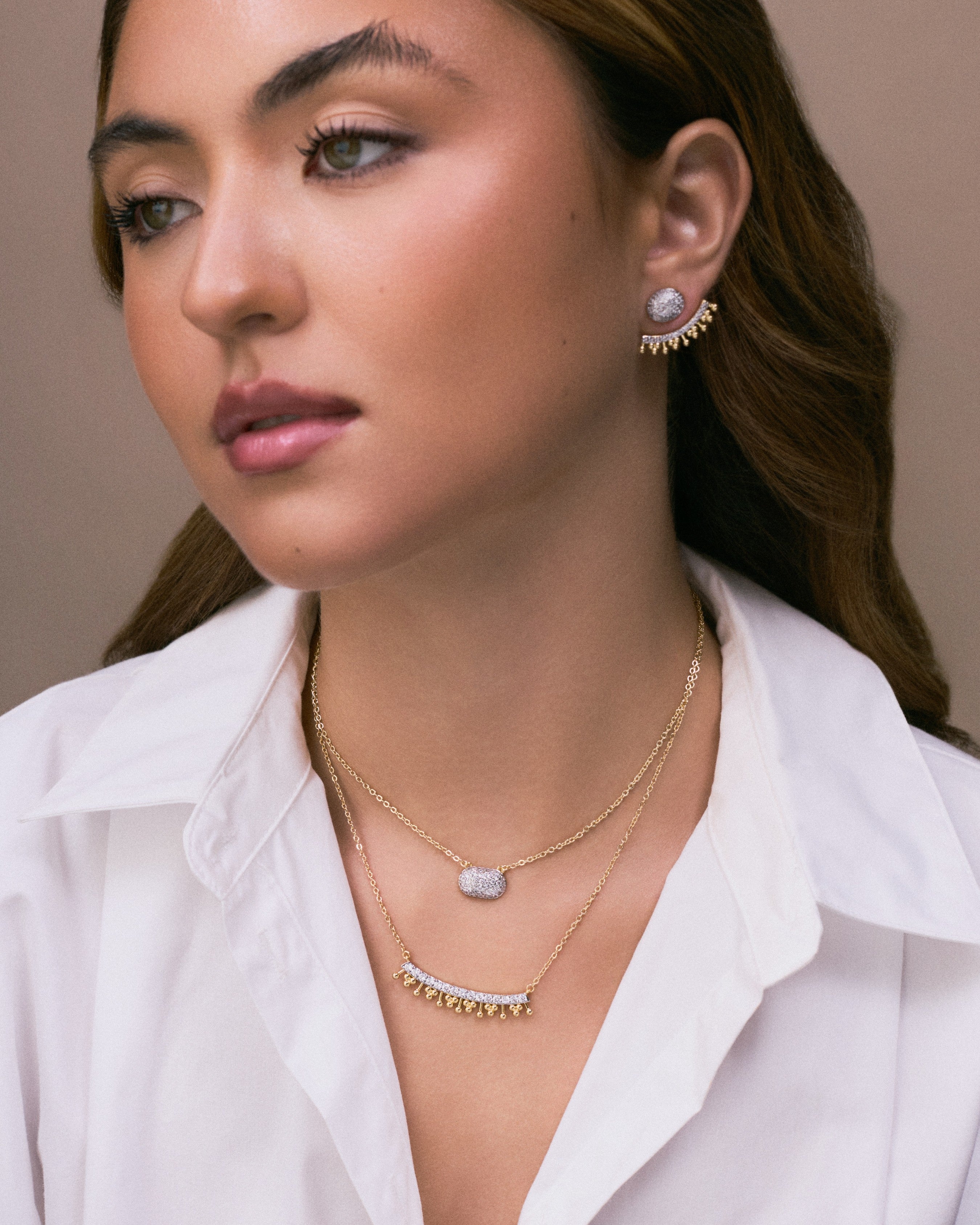 A close-up of a woman wearing layered gold necklaces and a matching gold crescent earring with a white stone, paired with a white collared shirt against a neutral beige background.